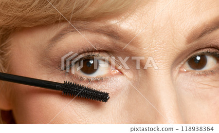 Woman applies mascara with precision, completing her makeup look, emphasizing attention to detail against white studio background. Woman applies mascara with precision, completing her makeup look, emphasizing attention to detail against white studio background. 118938364
