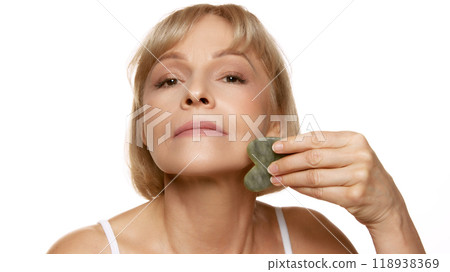 Woman uses scrapper, massaging chin, enhancing her skincare routine with natural beauty tools against white studio background. Woman uses scrapper, massaging chin, enhancing her skincare routine with natural beauty tools against white studio background. 118938369