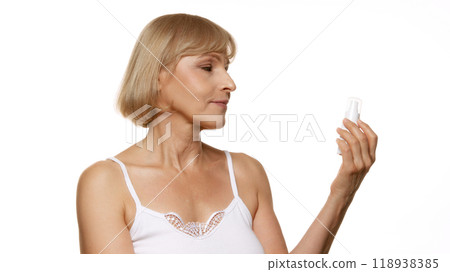 Inspecting her skincare product with calm gaze, beautiful woman chooses her self-care rituals with purpose against white studio background. 118938385