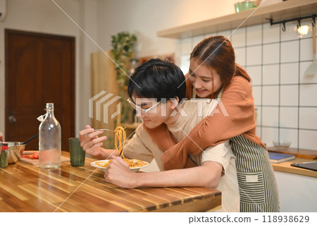 Pretty young woman embracing her boyfriend lovingly from behind while he eating pasta in the kitchen Pretty young woman embracing her boyfriend lovingly from behind while he eating pasta in the kitchen 118938629