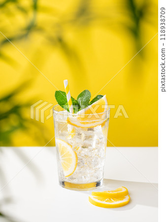 A glass of cold lemonade with lemon, mint and ice on a yellow and white background with a palm leaf. A glass of cold lemonade with lemon, mint and ice on a yellow and white background with a palm leaf. 118938709