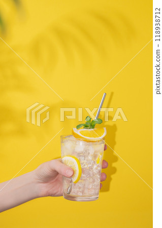A hand holds a glass of lemonade with lemon, ice and mint on a yellow background with shadow. 118938712