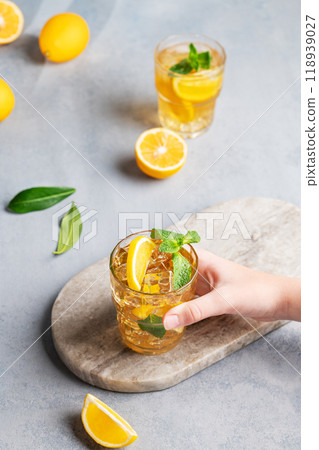 A hand takes a glass of iced tea with lemon and mint against a gray background with citrus fruits. 118939027
