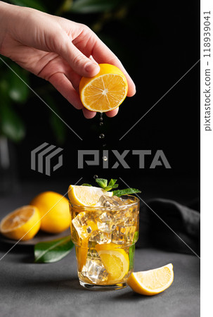 A hand squeezes juice from a lemon into a glass with lemonade with ice and mint on a dark background A hand squeezes juice from a lemon into a glass with lemonade with ice and mint on a dark background 118939041