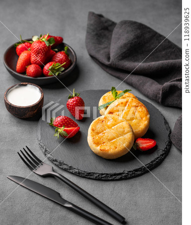 Cottage cheese, cheesecakes or syrniki with sour cream, strawberries and mint leaves on black slate Cottage cheese, cheesecakes or syrniki with sour cream, strawberries and mint leaves on black slate 118939625