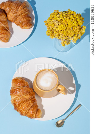 Flat lay of coffee cup and fresh croissants on blue background with yellow flowers and shadow. 118939635