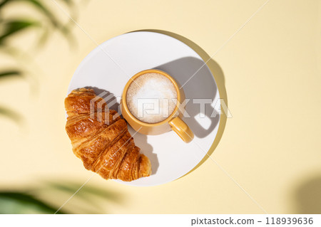 Flat lay of coffee cup and fresh croissant on a white plate on a yellow background with palm leaf Flat lay of coffee cup and fresh croissant on a white plate on a yellow background with palm leaf 118939636