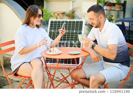 Man and woman charging smartphone from solar panel, relaxing in cafe. Cute couple using portable solar charger for devices. Charming couple sitting on terrace on background of solar battery. Man and woman charging smartphone from solar panel, relaxing in cafe. Cute couple using portable solar charger for devices. Charming couple sitting on terrace on background of solar battery. 118941190