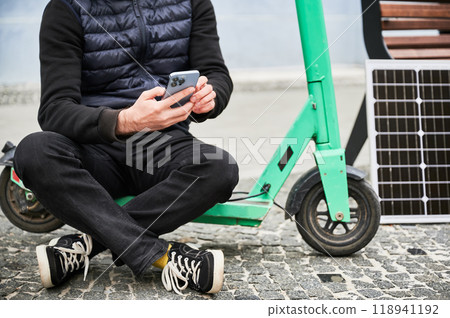 Cropped view of man sits on electric scooter with solar panel and smartphone while charging. Integration of renewable energy in personal transportation, highlighting sustainable urban lifestyle. 118941192