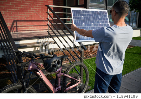 Back view of man holding solar panel toward the sun for charging electric mountain bike. Pink mountain bicycle with black helmet on handlebars. Scene set against brick building. 118941199