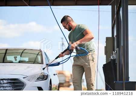 Washing luxury white auto with water gun on an open air car wash. Confident man cleaning his car with high pressure water jet. Technical service concept. 118941218