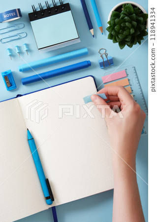 Top view of neat desk. Hand sticks a post-it note in an open notebook with a blank page. Around blue office supplies pens, pencils, felt-tip pens, sharpener, post-it notes. Copyspace. 118941334