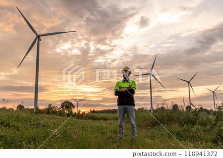 Professional Man Maintenance engineers working in wind turbine farm at sunset. Engineer Man standing among Wind Energy Turbine. Professional Man Maintenance engineers working in wind turbine farm at sunset. Engineer Man standing among Wind Energy Turbine. 118941372