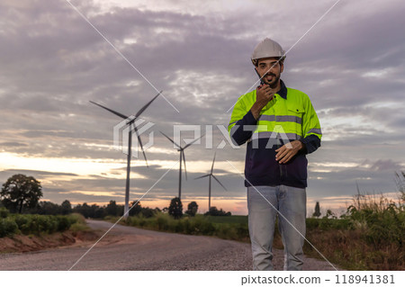 Professional Man Maintenance engineers working in wind turbine farm at sunset. Engineer Man standing among Wind Energy Turbine. Professional Man Maintenance engineers working in wind turbine farm at sunset. Engineer Man standing among Wind Energy Turbine. 118941381
