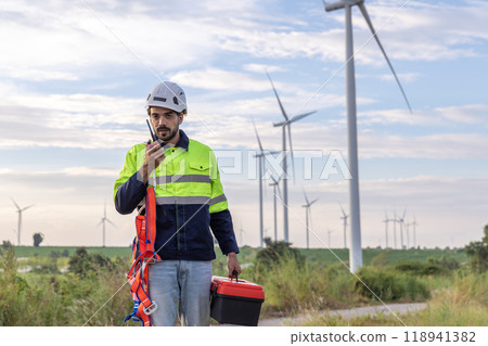 Professional Man Maintenance engineers working in wind turbine farm at sunset. Engineer Man standing among Wind Energy Turbine. Professional Man Maintenance engineers working in wind turbine farm at sunset. Engineer Man standing among Wind Energy Turbine. 118941382