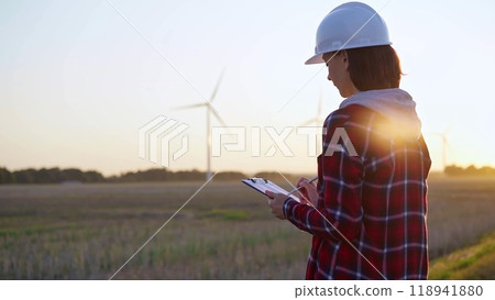 Adult woman engineer wearing white cask is taking notes on a clipboard on a field with wind turbines, as the sun sets, back view 118941880