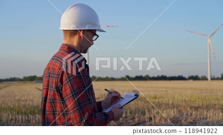 Man engineer, wearing a white protective helmet is taking notes with a clipboard in a field with wind turbines, as the sun sets. Clean energy and engineering concept 118941922