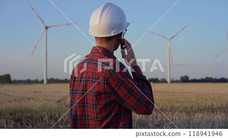Man engineer, wearing a white protective helmet is talking by smartphone in a field with wind turbines, as the sun sets. Clean energy and engineering concept 118941946