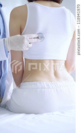 Doctor and patient. Physician wearing white medical gloves is using stethoscope examining woman lungs from the back side in clinic. Medicine and health care 118941970