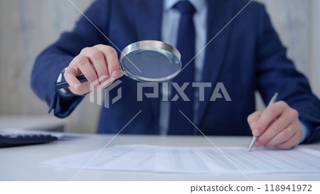 Businessman is carefully examining financial documents with a magnifying glass, highlighting the importance of thorough analysis in business. Audit and taxes 118941972