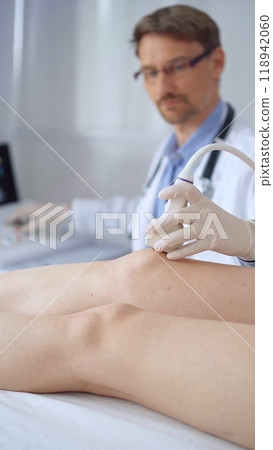Doctor man with blue protective gloves using ultrasound equipment on the knee of a female patient, analyzing the results on a monitor in the background. Medicine Doctor man with blue protective gloves using ultrasound equipment on the knee of a female patient, analyzing the results on a monitor in the background. Medicine 118942060