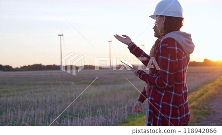 Man and woman, both engineers wearing white protective helmets, are checking and discussing something in a field with wind turbines as the sun sets. Clean energy and engineering concept Man and woman, both engineers wearing white protective helmets, are checking and discussing something in a field with wind turbines as the sun sets. Clean energy and engineering concept 118942066