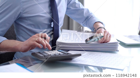 Businessman in blue short is using calculator and reviewing ring folder of financial documents with magnifying glass at desk in modern office. Audit and taxes in business concept 118942148