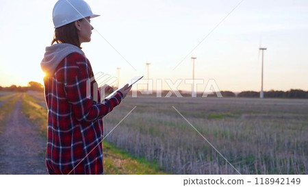 Woman engineer wearing a white protective helmet is taking notes with a tablet computer on a field with wind turbines, as the sun sets. Clean energy and engineering concept 118942149