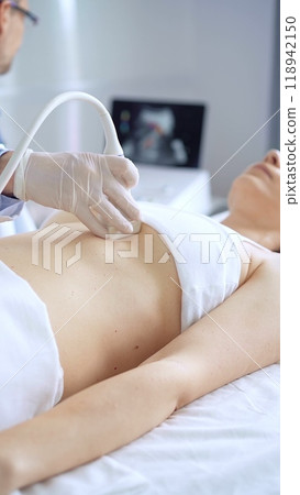 Doctor man wearing white protective gloves is using ultrasound equipment on the breast of a female patient. Mammary gland health 118942150
