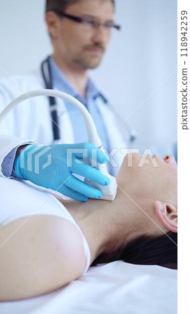 Doctor man wearing blue protective gloves is using ultrasound machine to examine neck of female patient in clinic. Medicine, thyroid gland examination 118942259