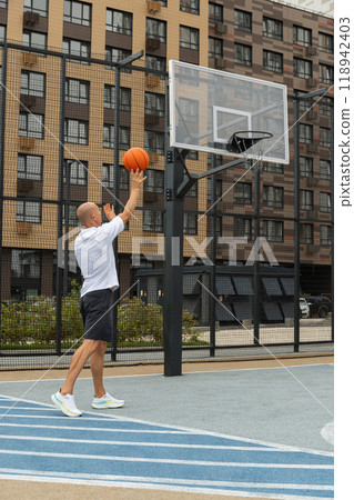 Sporty man tries to throw ball into basketball hoop on sports court at courtyard. Street style. Game moment. Sporty man tries to throw ball into basketball hoop on sports court at courtyard. Street style. Game moment. 118942403