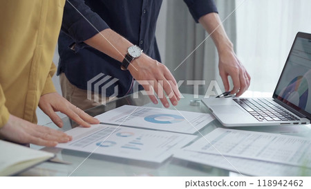 Business people, professionals analyzing financial charts and data on a desk while standing and using laptop computer in office. Dark blue shirt and yellow blouse Business people, professionals analyzing financial charts and data on a desk while standing and using laptop computer in office. Dark blue shirt and yellow blouse 118942462