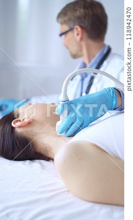Doctor man wearing blue protective gloves is using ultrasound equipment on the neck of a patient woman, lying for examination. Medicine concept 118942470