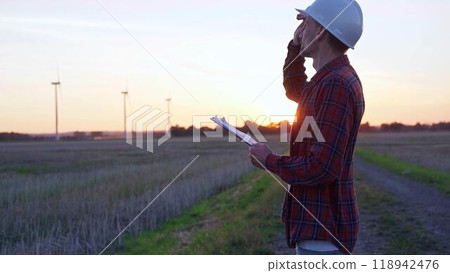 Man engineer wearing a white protective helmet is taking notes with a clipboard in a field with wind turbines, as the sun sets. Clean energy and engineering concept 118942476