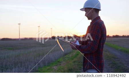 Man engineer wearing a white protective helmet is taking notes with a clipboard in a field with wind turbines, as the sun sets. Clean energy and engineering concept Man engineer wearing a white protective helmet is taking notes with a clipboard in a field with wind turbines, as the sun sets. Clean energy and engineering concept 118942492