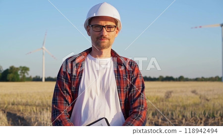 Man engineer at work is standing straight and taking notes with a clipboard in a field with wind turbines, as the sun sets. Clean energy concept Man engineer at work is standing straight and taking notes with a clipboard in a field with wind turbines, as the sun sets. Clean energy concept 118942494