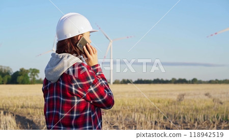 Woman engineer is using a smartphone in a field with wind turbines as the sun sets. Concept of clean energy and engineering audit Woman engineer is using a smartphone in a field with wind turbines as the sun sets. Concept of clean energy and engineering audit 118942519