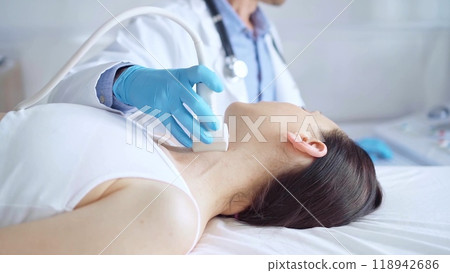 Doctor man wearing blue medical gloves is using ultrasound equipment on neck of female patient lying down for a medical examination Doctor man wearing blue medical gloves is using ultrasound equipment on neck of female patient lying down for a medical examination 118942686