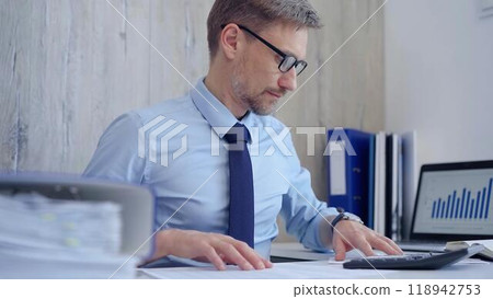 Male accountant with blue t-shirt concentrating while calculating costs and taking notes at his desk. Taxes, audit in business 118942753