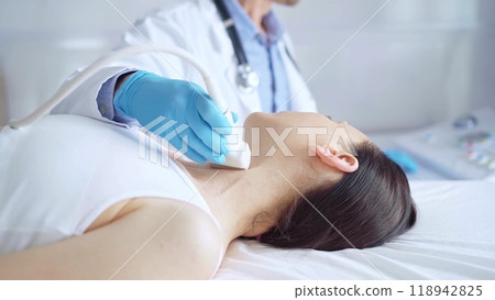 Doctor man wearing blue medical gloves is using ultrasound equipment on neck of female patient lying down for a medical examination 118942825