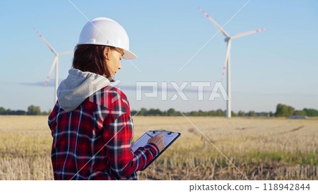 Woman engineer wearing a white protective helmet is taking notes with a clipboard in a field with wind turbines, as the sun sets. Clean energy and engineering concept Woman engineer wearing a white protective helmet is taking notes with a clipboard in a field with wind turbines, as the sun sets. Clean energy and engineering concept 118942844