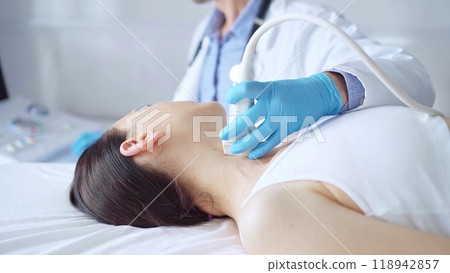 Doctor man wearing blue medical gloves is using ultrasound equipment on neck of female patient lying down for a medical examination 118942857
