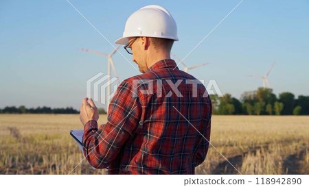 Man engineer at work is wearing a white protective helmet and taking notes with a clipboard in a field with wind turbines, as the sun sets. Clean energy concept 118942890