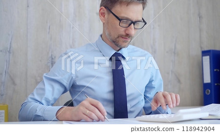 Male accountant with blue t-shirt concentrating while calculating costs and taking notes at his desk. Taxes, audit in business Male accountant with blue t-shirt concentrating while calculating costs and taking notes at his desk. Taxes, audit in business 118942904