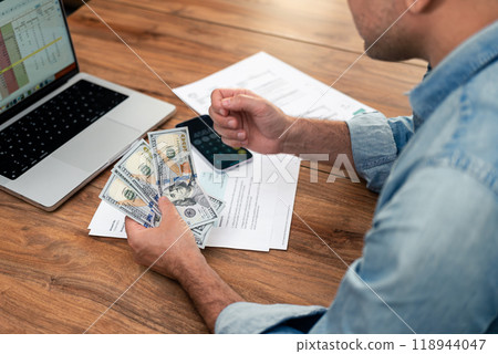 Close-up of a man holding dollar bills while working on household expenses at a desk. Financial planning and budgeting with utility bills and a laptop. Home finance and payment management concept Close-up of a man holding dollar bills while working on household expenses at a desk. Financial planning and budgeting with utility bills and a laptop. Home finance and payment management concept 118944047