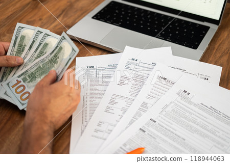 Hands counting hundred-dollar bills with tax forms and a laptop on a wooden table. Financial management and tax filing concept. 118944063
