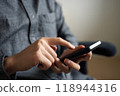 A hand operating a black smartphone. A person sitting on a chair tapping on the touch screen of a smartphone 118944316