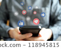 A black smartphone is held in both hands with floating icons representing likes, bookmarks and comments 118945815