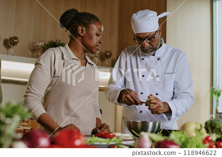 Low angle shot of Indian chef peeling avocado to add it into salad while African American female intern watching his actions 118946622