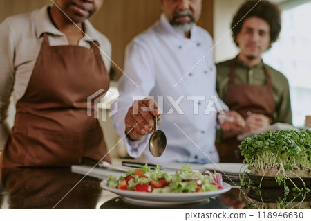 Medium close up of chefs hand holding spoon and adding olive oil into salad while his assistants standing next to him 118946630